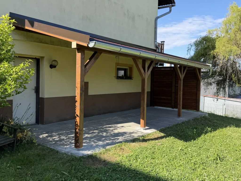 Dokončená dřevěná pergola se střechou z červeného asfaltového šindele navazující na dům
